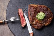 Medium gebratenes Steak mit Kräuterbutter, mit Messer und Gabel angeschnitten auf neumodischer, runder Schieferplatte serviert Medium gebratenes Steak mit Kräuterbutter, mit Messer und Gabel angeschnitten auf neumodischer, runder Schieferplatte serviert